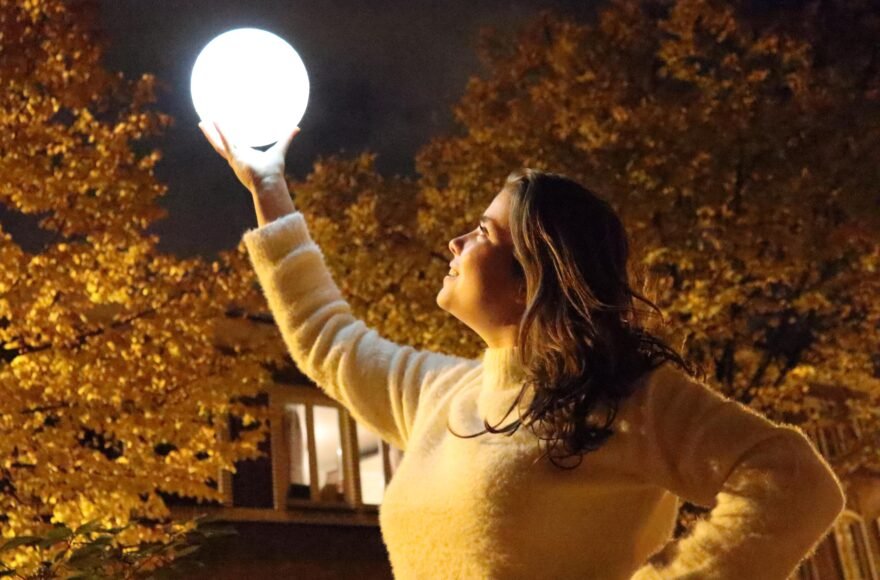 Je ziet een meisje met bruin haar en een witte trui op straat staan. In de straat is het donker en je ziet bomen met gele bladeren. Het meisje houdt 1 arm omhoog en daarin zit een lichtgevende bol. Het meisje kijk er naar en lacht