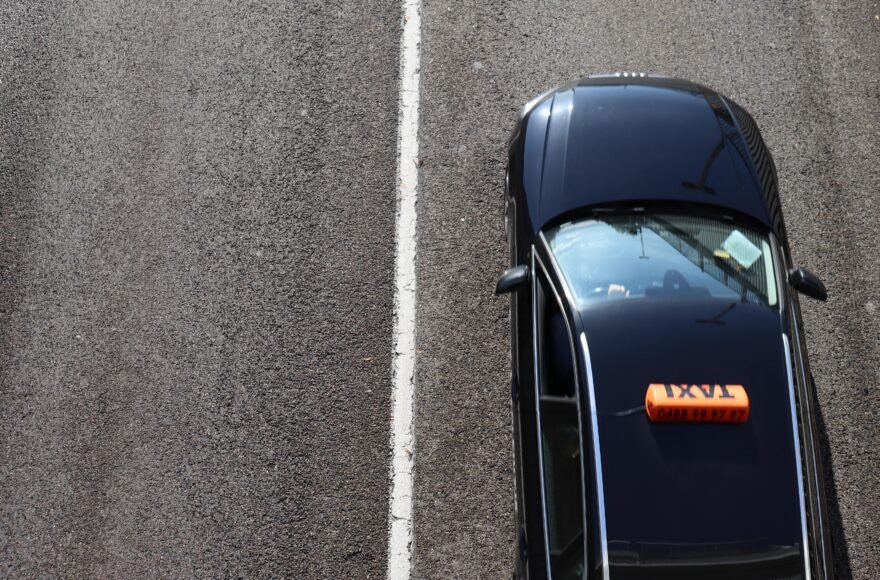 Een taxi rijdt op de baan. Het linkerbaanvak is leeg, dan is er een witte streep en op het rechterbaanvak rijdt een zwarte taxi.